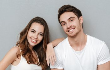 A smiling man and woman in white shirts stand against a gray background, with the woman's arm resting on the man's shoulder.