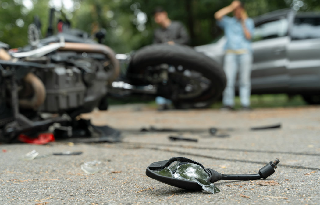 A damaged motorcycle and broken mirror lie on the road after an accident, with two people and a car visible in the blurred background.