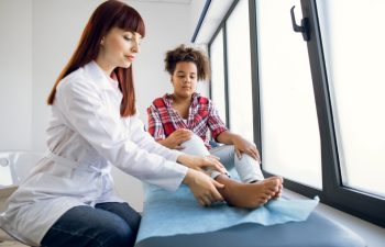 Trauma on the child's leg. Female Caucasian doctor orthopedist, examining foot of pretty mixed race teen girl in clinic. Child suffering from pain at doctor's office